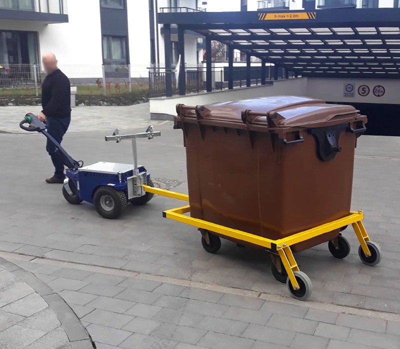 Powered tug towing a large waste bin on a yellow wheeled trolley