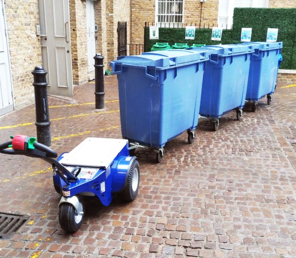 Powered tug transporting a connected train of commercial waste bins